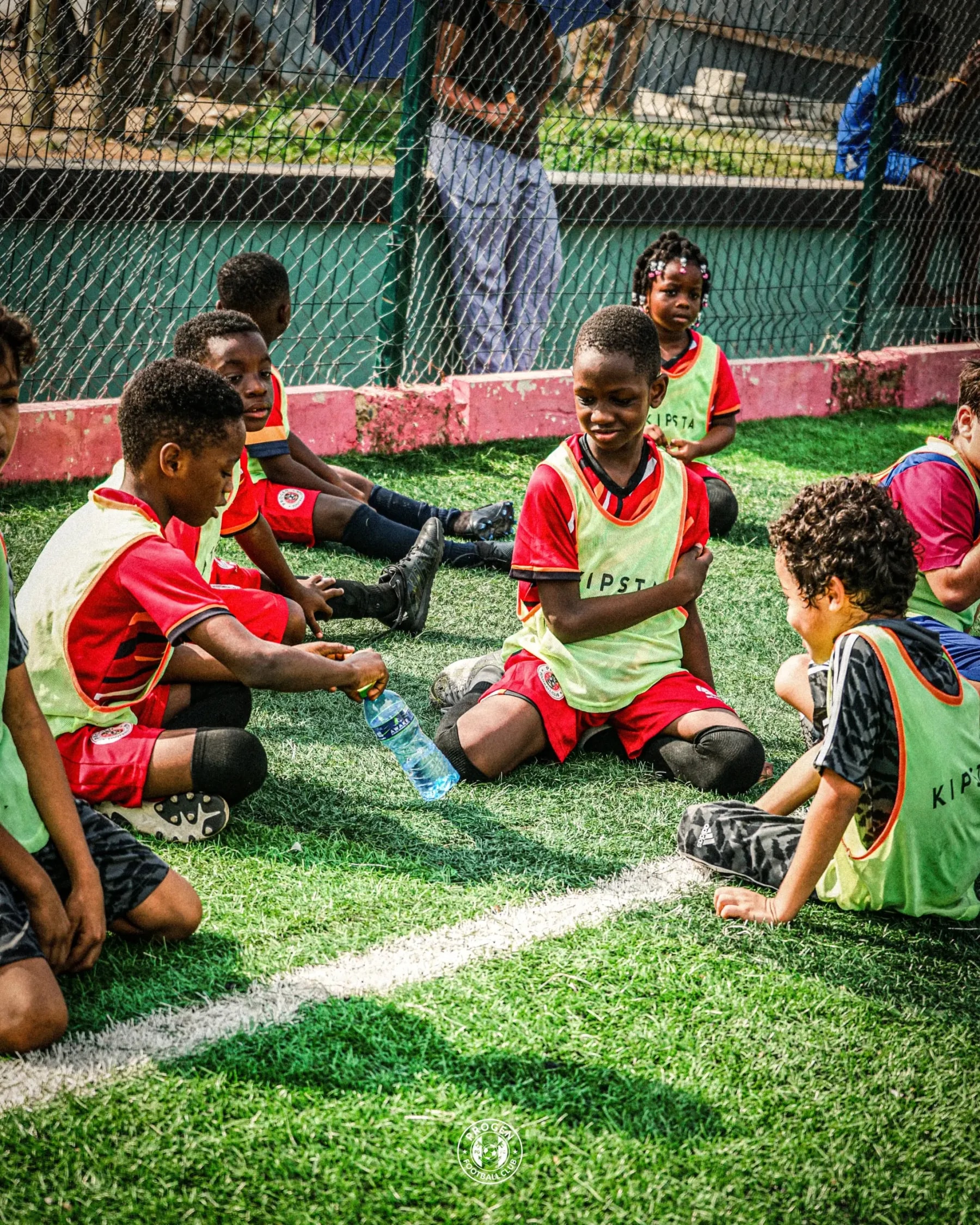 Academy players in training bibs sitting on the pitch watching a bottle-flip challenge